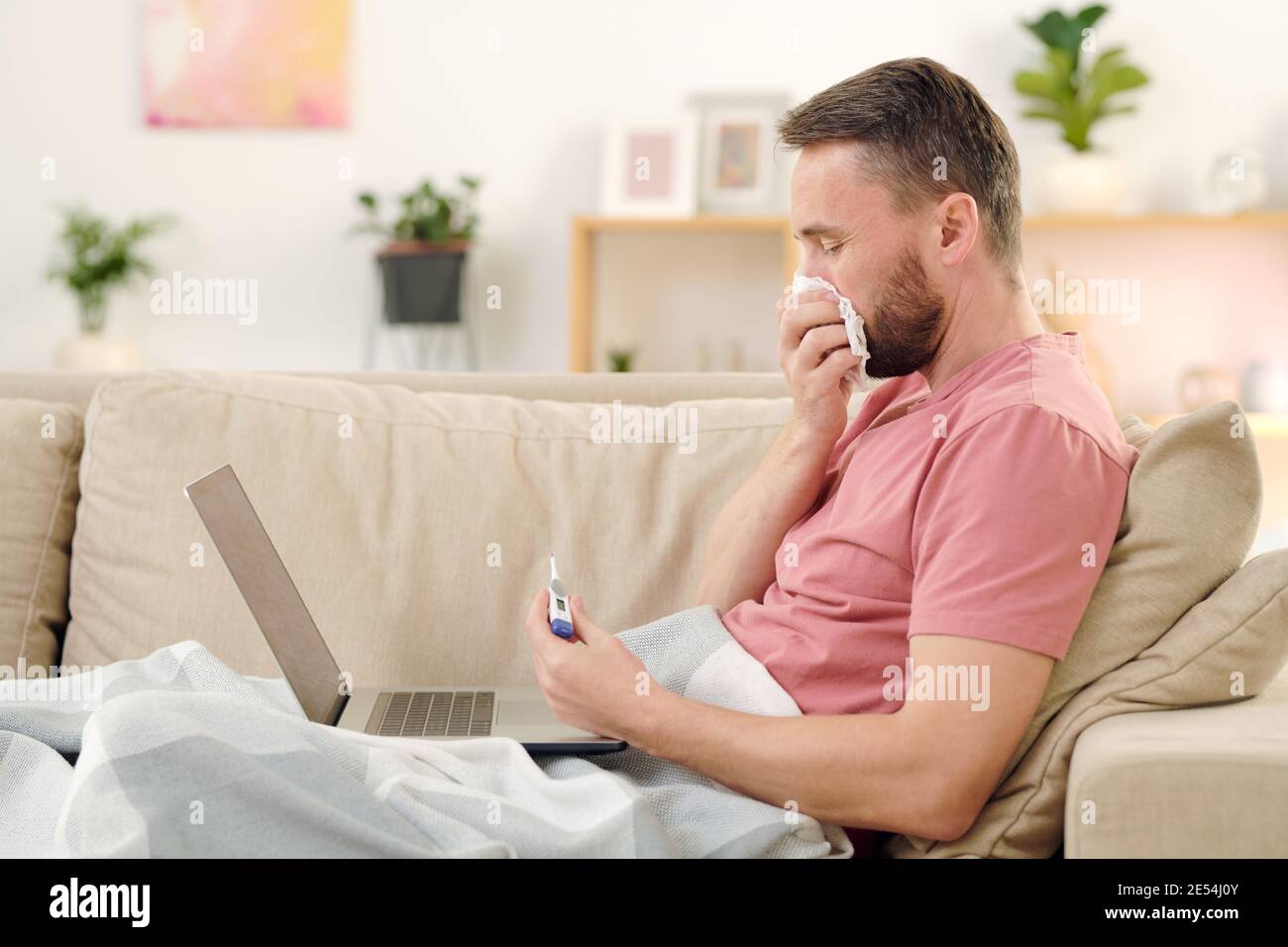 Seitenansicht des jungen kranken Mannes, der Thermometer und ansieht Er bläst seine Nase, während er auf der Couch unter der Decke sitzt Beratung Online-Arzt Stockfoto
