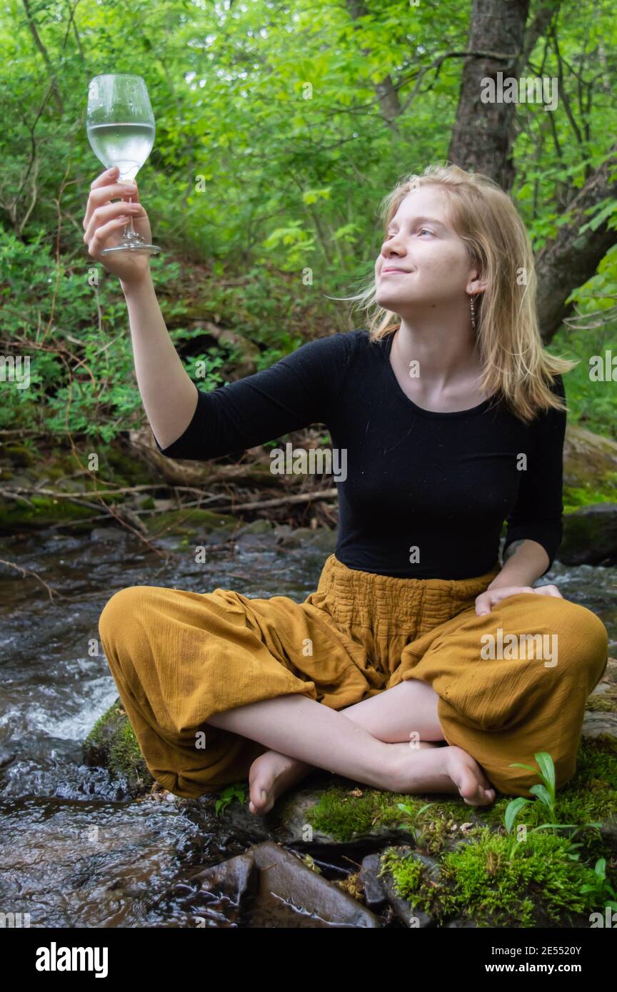 Porträt einer jungen lächelnden Frau, die mit gekreuzten Beinen auf einem moosbedeckten Felsen am Ufer eines Flusses sitzt und ein Weinglas betrachtet. Stockfoto