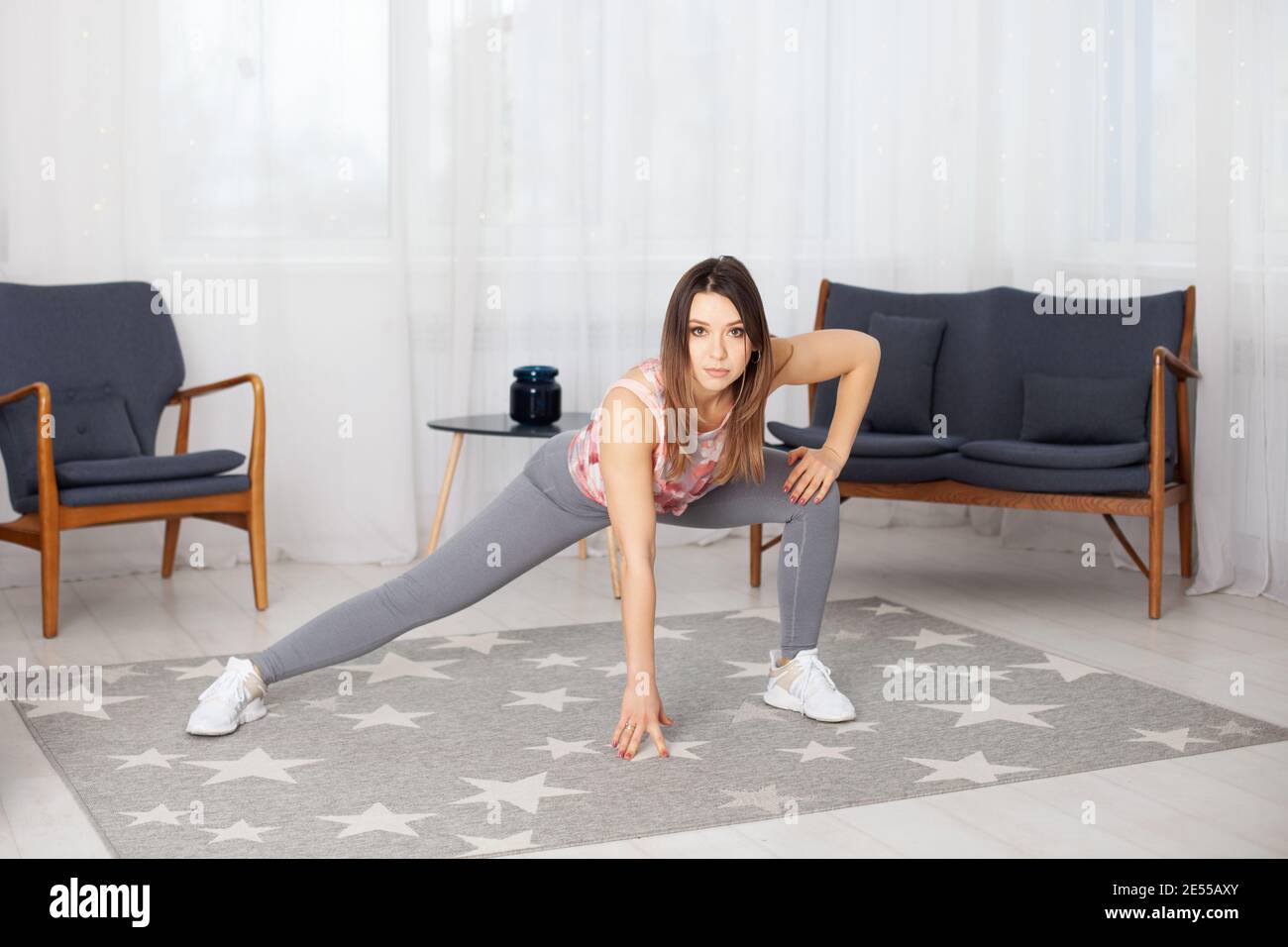 Durchgehende junge Frau in grauen Leggings hockt seitlich auf einem Bein in einem Heim-Interieur Stockfoto