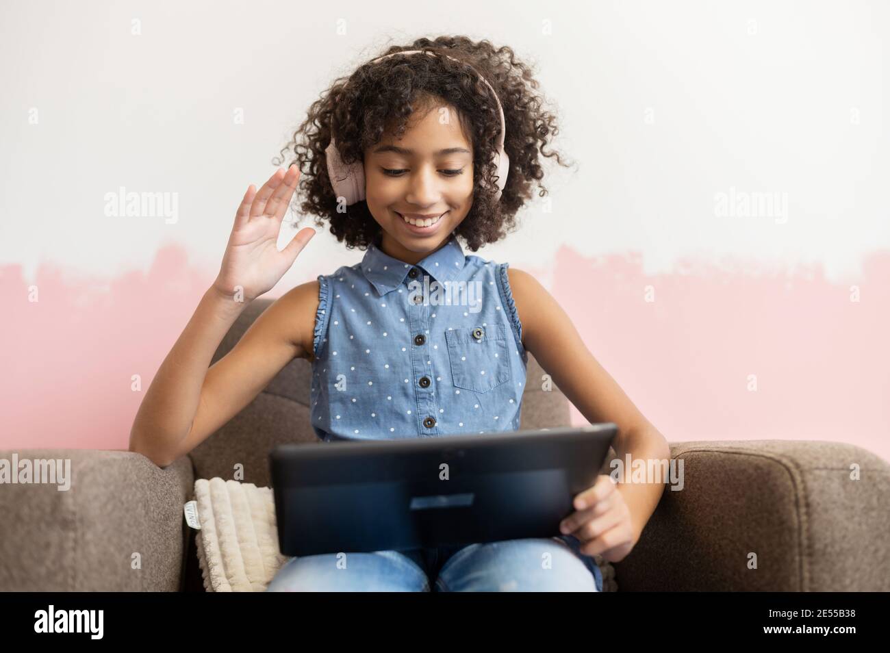 Fröhliches afrikanisches Schulmädchen, das zu Hause mit einem digitalen Tablet am Sessel sitzt, in die Webcam winkt und online mit Freunden chattet Stockfoto
