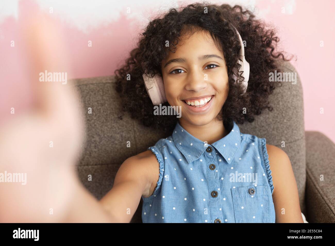 Ein fröhliches, lockiges afroamerikanisches Mädchen, das einen Kopfhörer trägt, nimmt ein Selfie auf. Ein Nahaufnahme Porträt von ziemlich biracial Schule Alter Mädchen blogger Stockfoto