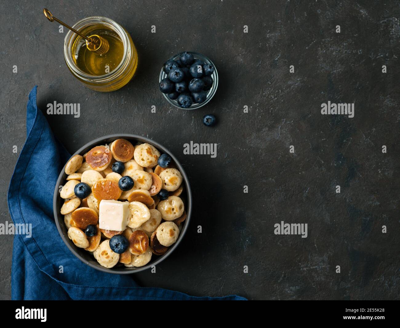 Trendiges Essen: Pfannkuchen, Müsli, Butter und Heidelbeeren. Haufen Mini-Cerealien-Pfannkuchen in Boul auf dunklem Hintergrund. Platz rechts für Text kopieren. Ansicht von oben oder flach liegend. Stockfoto