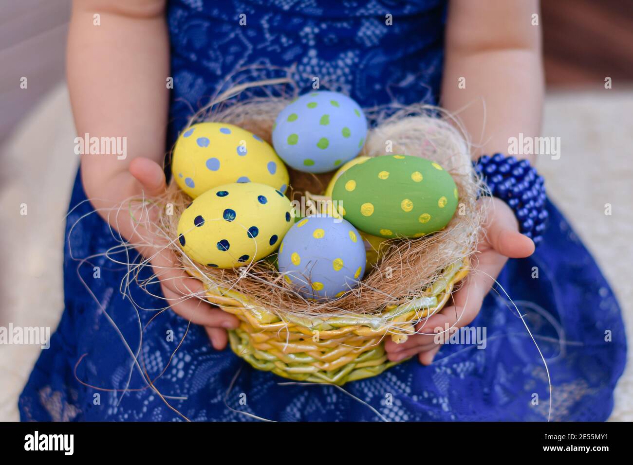 Ein Korb mit bunten Ostereiern in den Händen eines kleinen Mädchens in einem blauen Kleid. Stockfoto