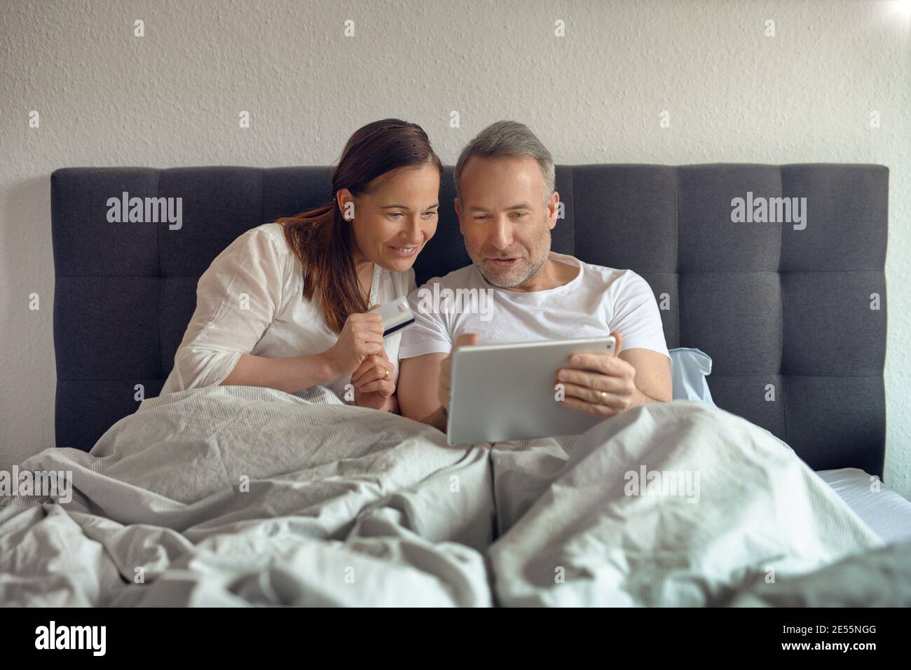 Ein glückliches Paar mittleren Alters, das morgens im Bett saß und trank Kaffee und Online-Shopping mit einem digitalen Tablet Stockfoto
