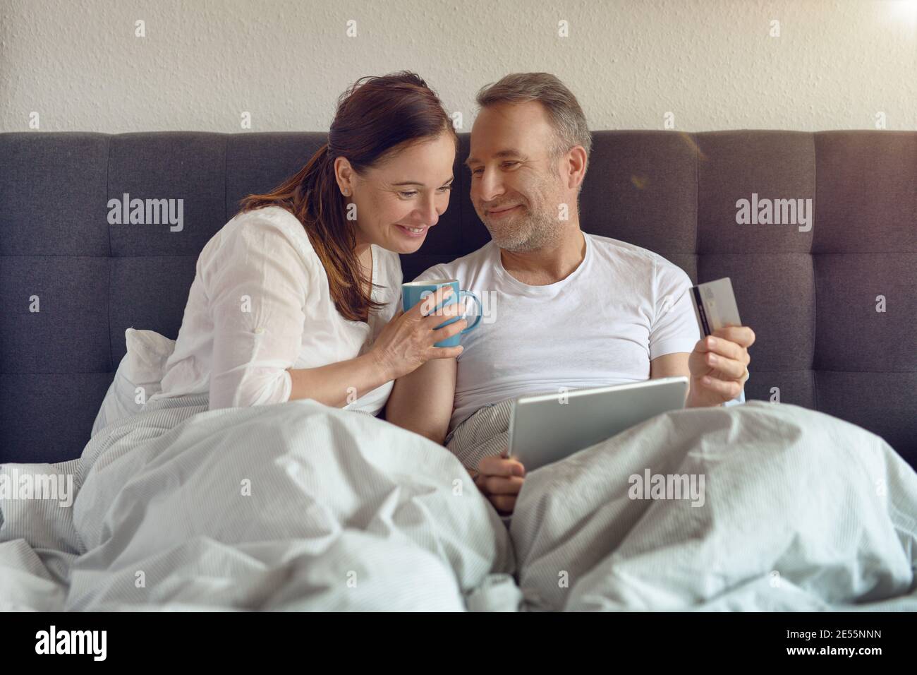 Ein glückliches Paar mittleren Alters, das morgens im Bett saß und trank Kaffee und Online-Shopping mit einem digitalen Tablet Stockfoto