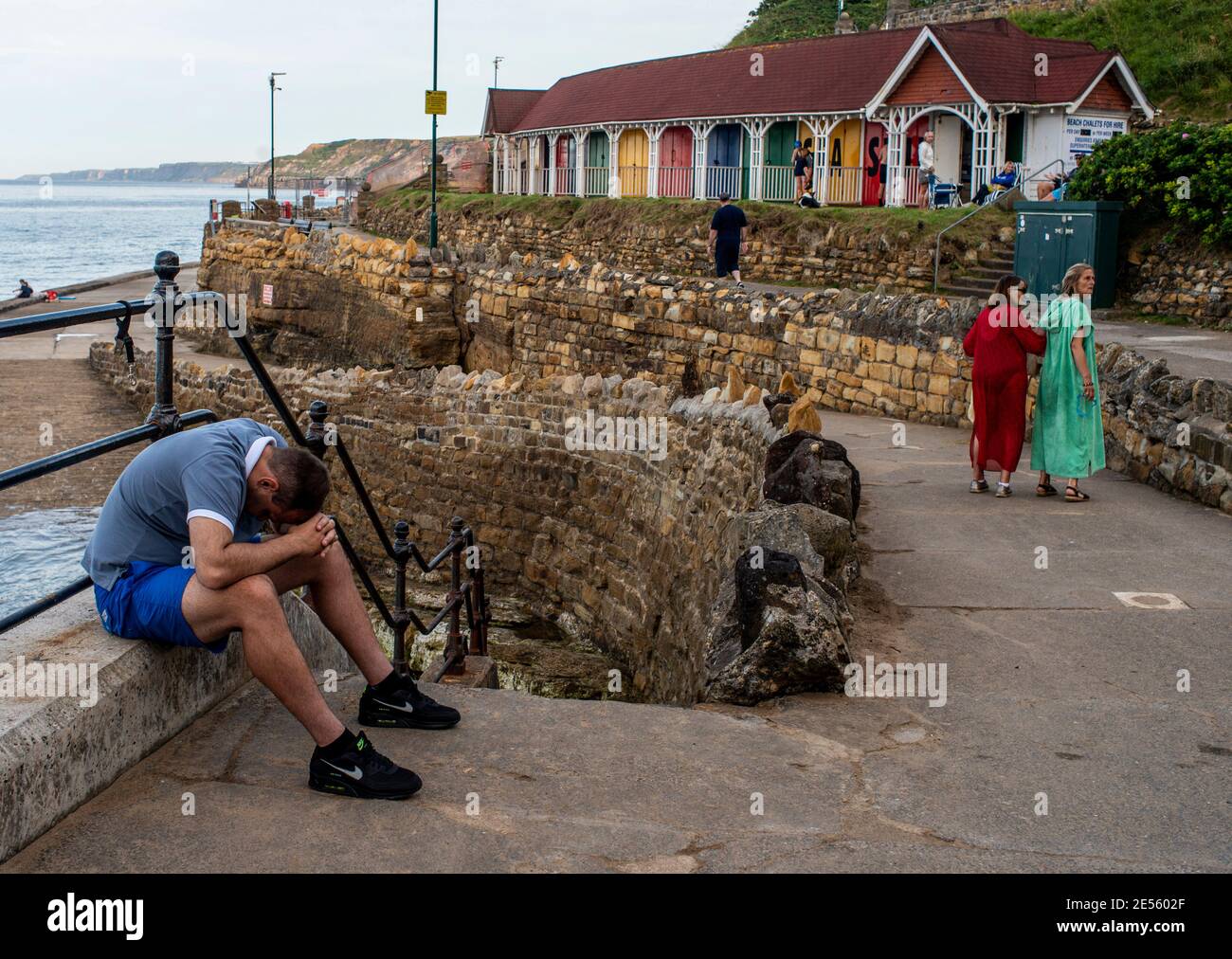 Ein Mann, der mit dem Kopf auf Händen und Knien am Scarborough Beach sitzt, während 2 Damen in hellen Kleidern im Hintergrund eher verloren aussehen. Stockfoto