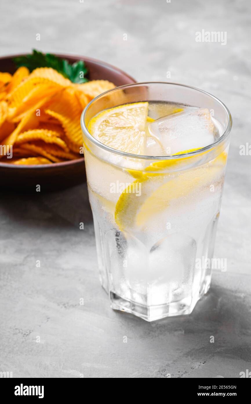 Frisches eiskaltes Wasser Getränk mit Zitrone in der Nähe von gebratenen gewellten goldenen Kartoffelchips mit Petersilienblatt in Holzschüssel auf Beton Hintergrund, Winkelansicht Stockfoto