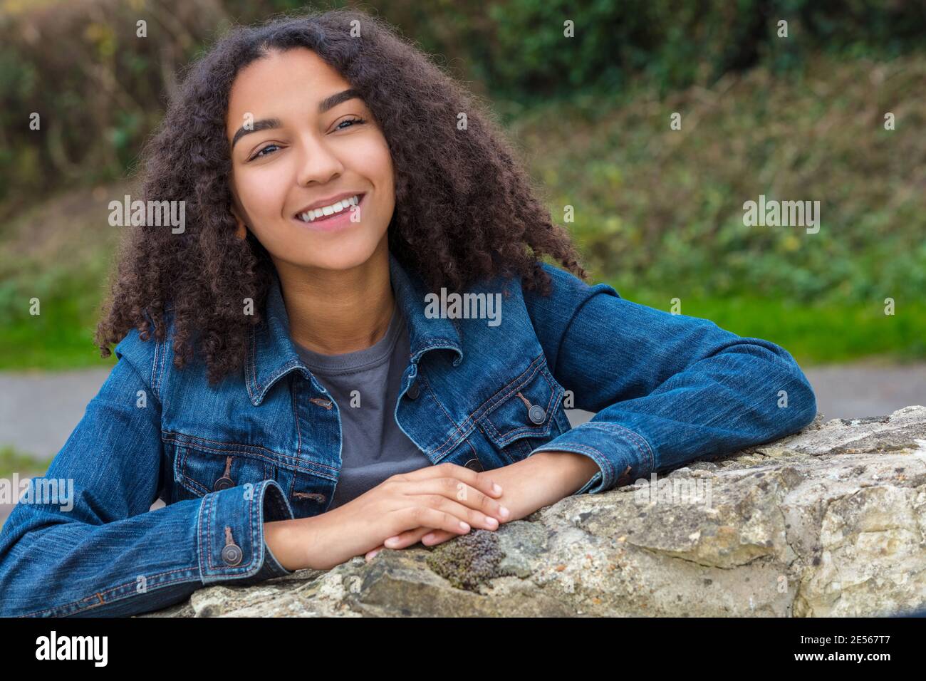 Outdoor-Porträt von schönen glücklich gemischte Rasse afroamerikanische Mädchen Teenager weibliches Kind lächelnd mit perfekten Zähnen tragen Denim Jacke Sich darauf stützen Stockfoto