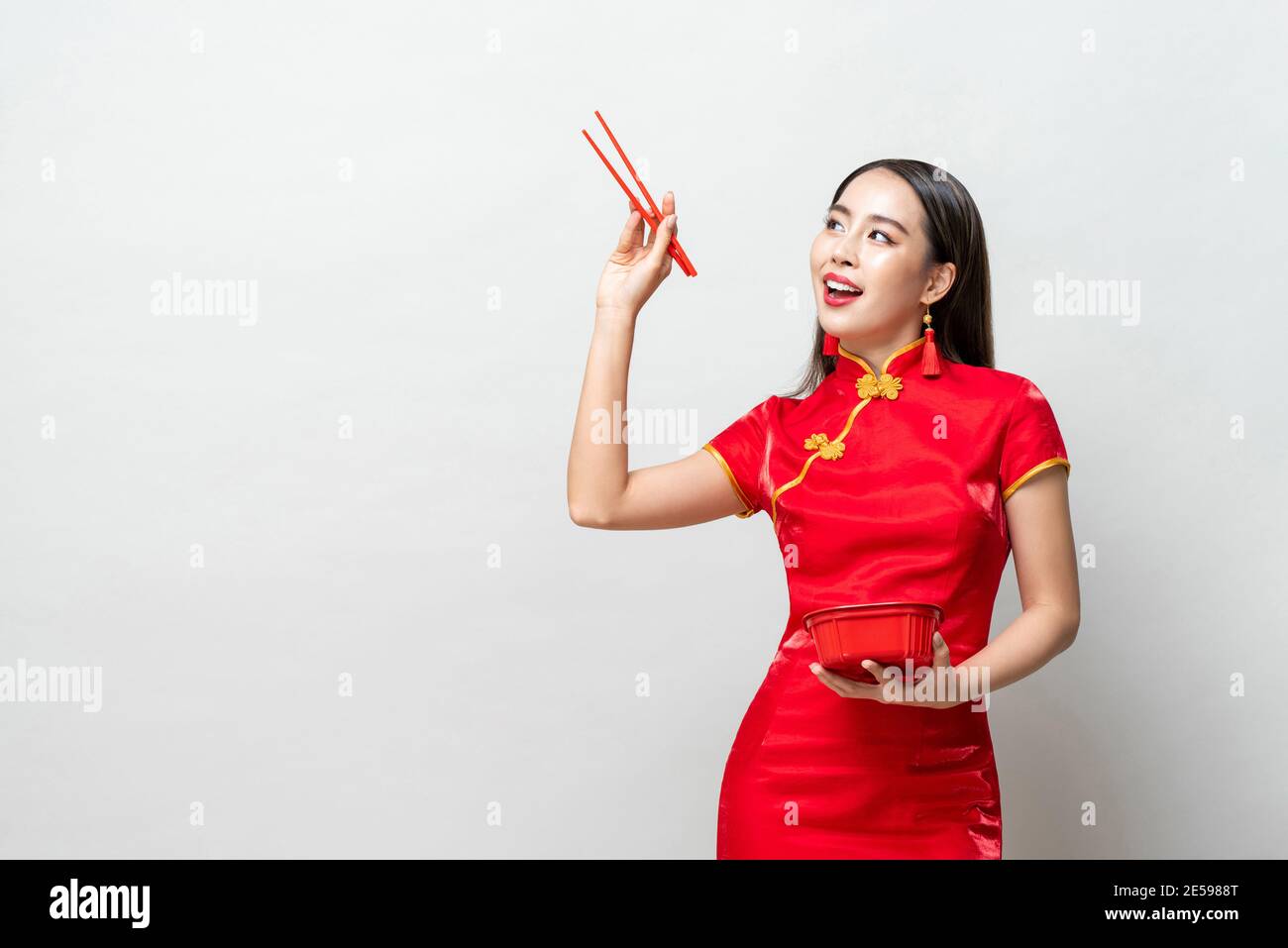 Asiatische Frau in traditionellen roten chinesischen Qipao Kleid hält Kunststoff Lebensmittelkarton und Essstäbchen, die nach oben schauen, um Platz zu schaffen studio isoliert hellgrau Stockfoto