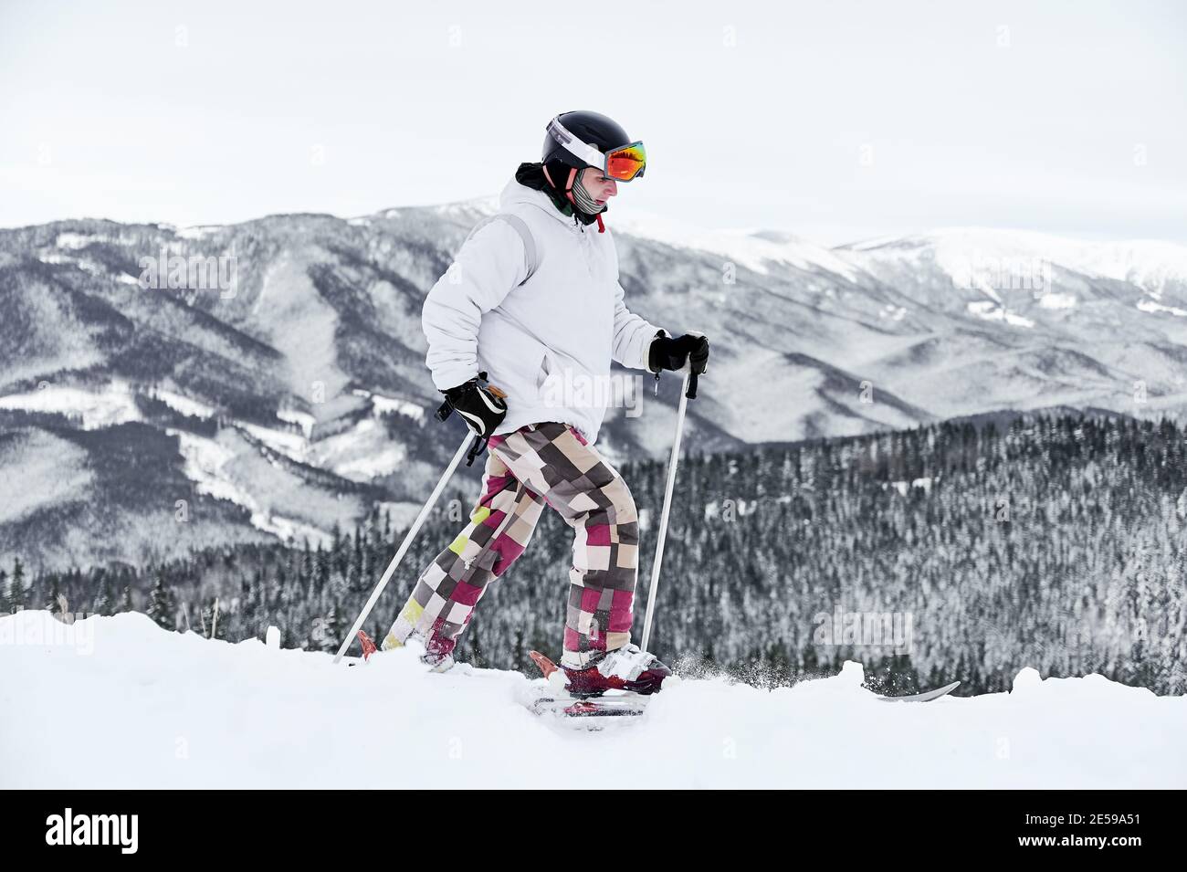 Horizontale Momentaufnahme des jungen Skifahrers in buntem Kostüm, der entlang verschneite Hügel gegen erstaunliche Berge auf Hintergrund läuft, Skitour. Seitenansicht, Kopierbereich, volle Länge Stockfoto