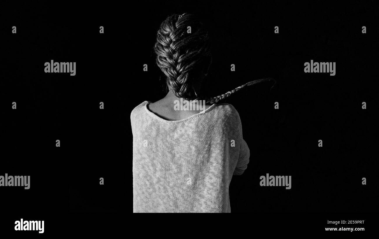 Ein Mädchen in einem weißen Strickpullover steht mit dem Rücken auf schwarzem Hintergrund. Schwarzweiß-Foto, Mitte. Stockfoto