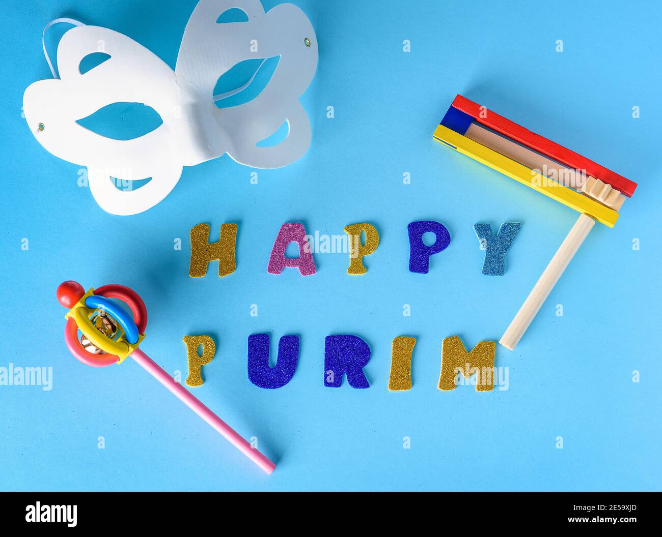 Happy Purim jüdischen Feier Feiertag Grußkarte. Weiße Karnevalsmaske, bunte hölzerne Noisemakers oder Grager auf blauem Hintergrund.Happy Purim Glitzer Text.Flat Lay. Stockfoto