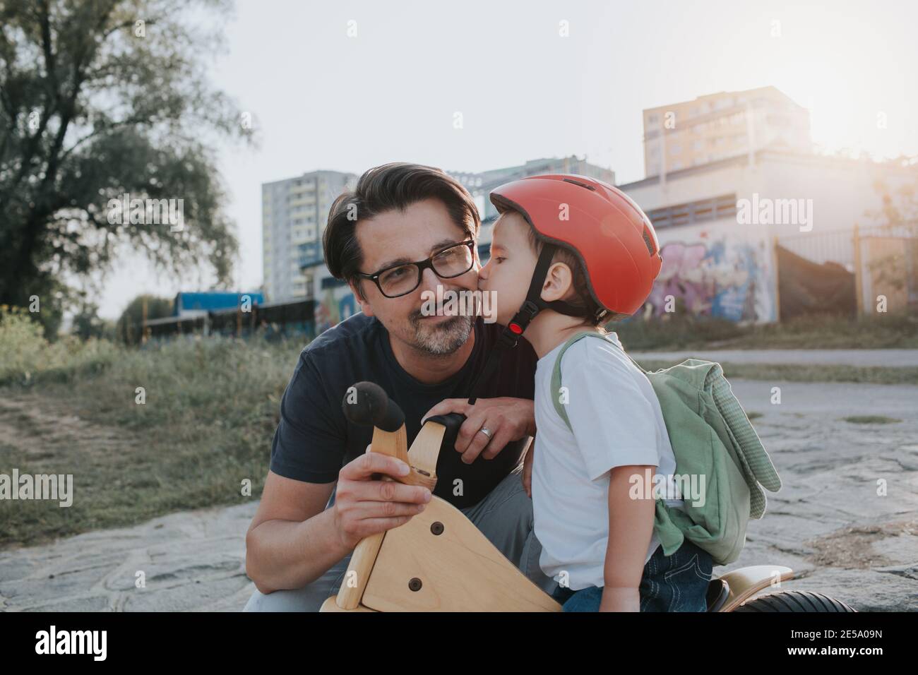 Lächelnder Vater bekommt einen Kuss von seinem kleinen Sohn auf einem Fahrrad. Kleiner Kleinkind Junge trägt einen Helm küssen seinen Papa während eines Abendspaziergangs. Stockfoto