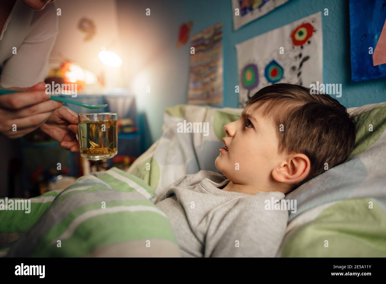 Ein krankes Kind, das in seinem Zimmer im Bett liegt und sich weigert, eine Medizin einzunehmen. Ein Junge, der nicht bereit ist, eine Arznei von seiner Mutter zu nehmen. Stockfoto
