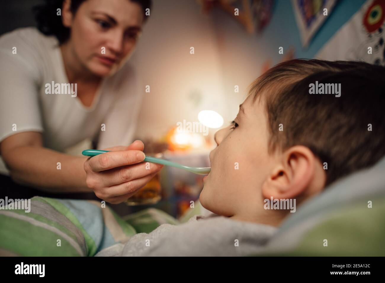 Ein krankes Kind liegt im Bett und nimmt eine Medizin von seiner Mutter. Eine Mutter, die ihrem kranken Sohn Medizin auf einem Löffel anbietet. Stockfoto