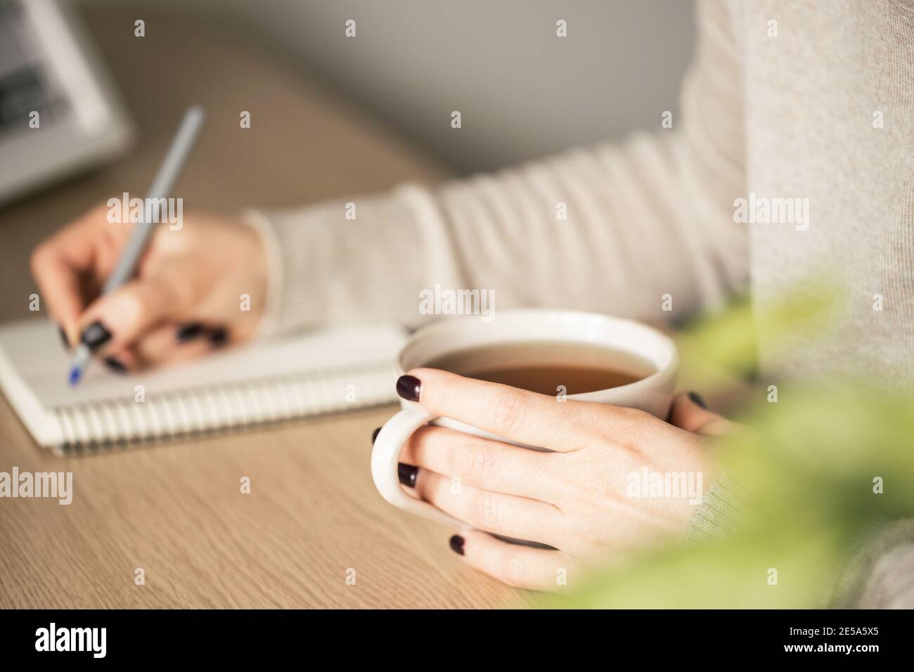 Frau in beige Pullover hält eine Tasse Teain eine Hand und sie schreibt. Selektiver Fokus. Stockfoto