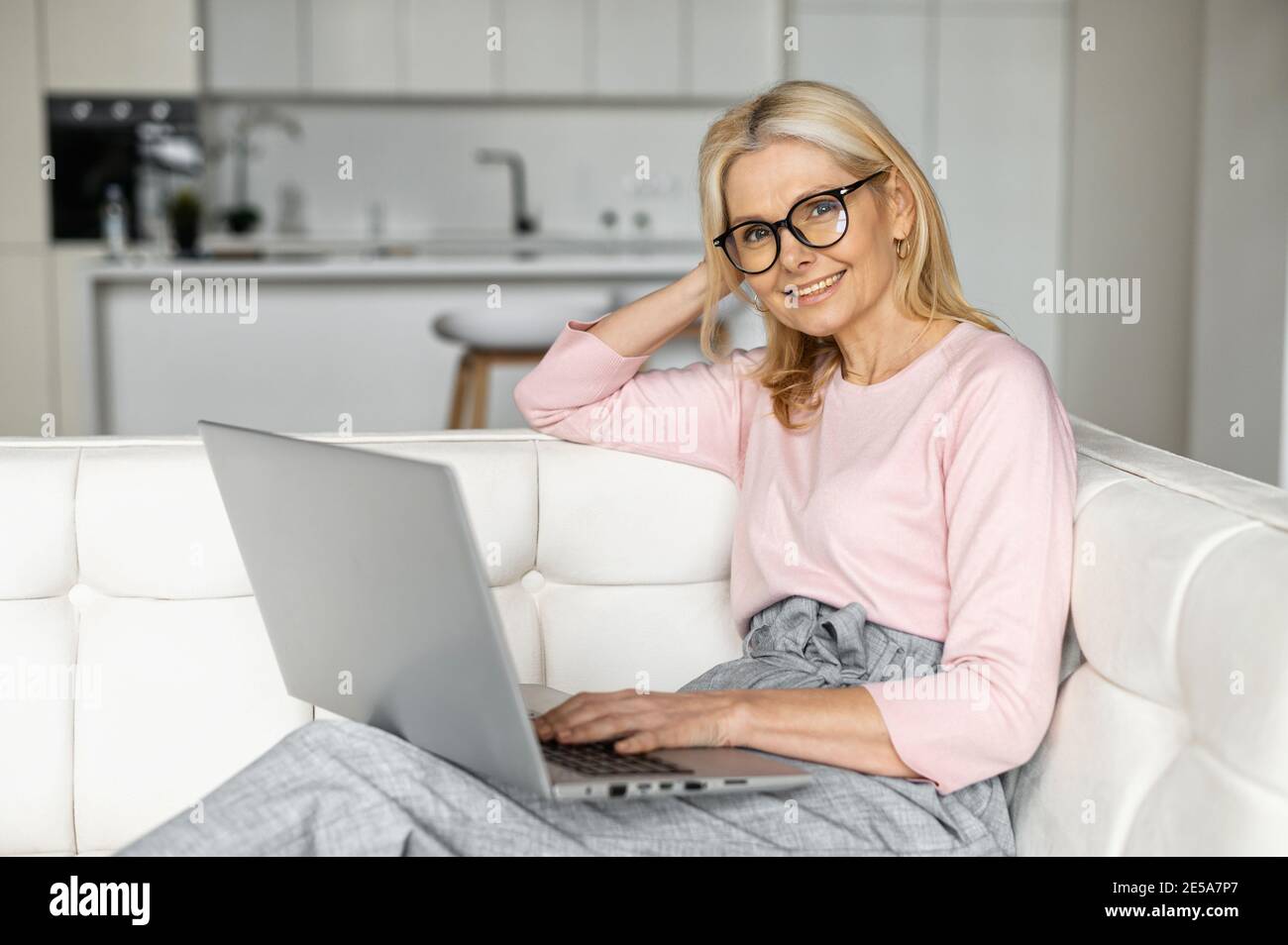 Entspannte Geschäftsfrau mittleren Alters, die auf der Couch sitzt und online auf dem Laptop chattet und E-Mails beantwortet. Eine fröhliche Dame mit Brille verbringen Stockfoto