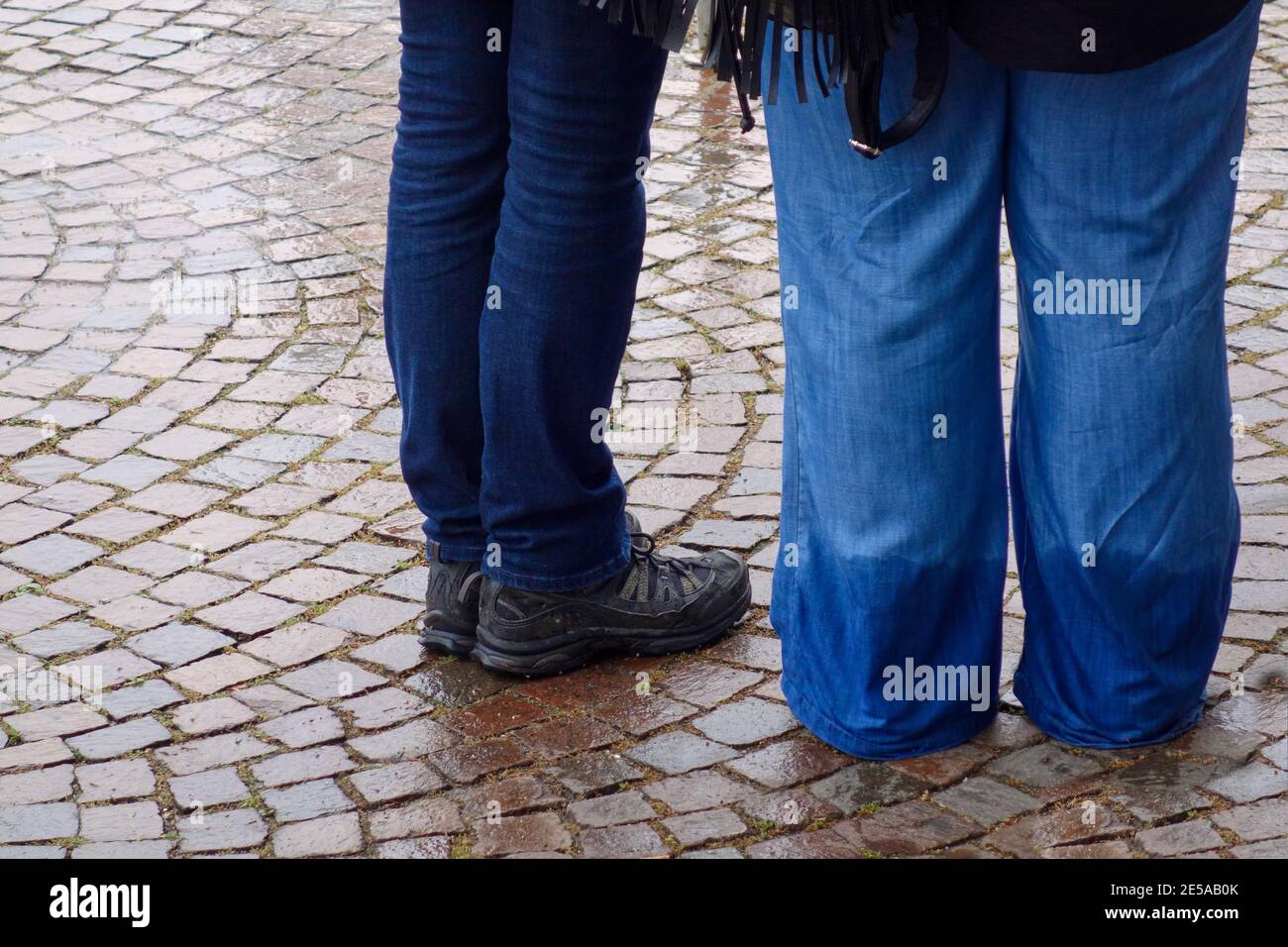 Das Bild zeigt die Beine von 2 Personen, die draußen im Nassen stehen, 1 die sehr nasse, regnernassgetränkte Jeans trägt Stockfoto