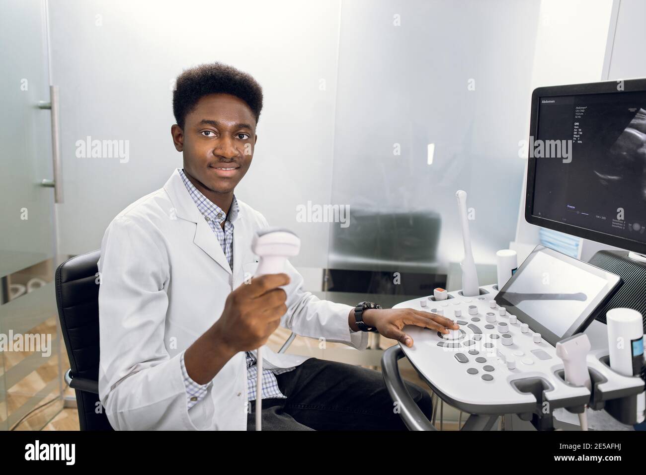 Lächelnder afrikanischer Mann Arzt mit Ultraschallaufnehmer in der Hand, der an einem modernen Ultraschallgerät im hellen Raum in der Klinik arbeitet. 4D-Hochformat Stockfoto