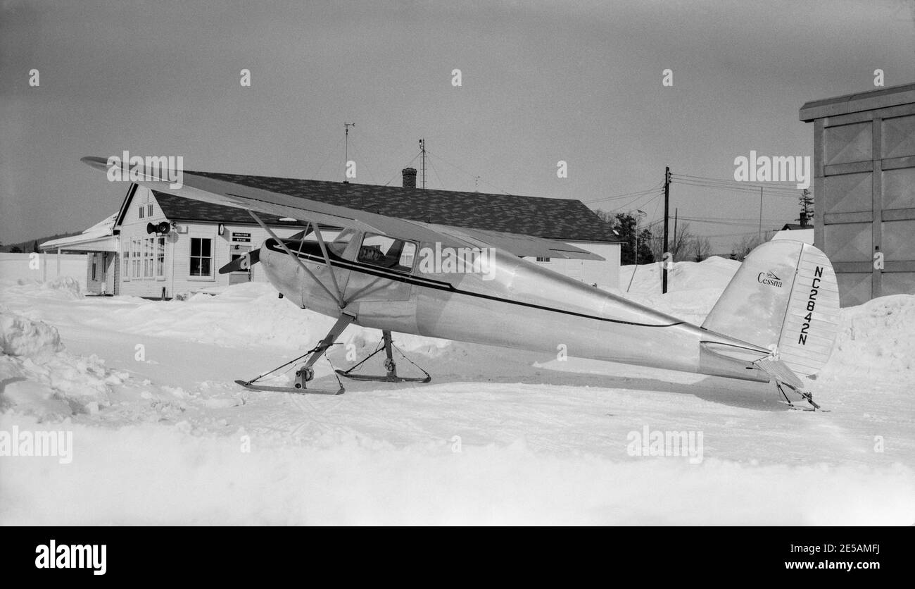 Vintage-amerikanische Schwarz-Weiß-Fotografie eines leichten Cessna 120-Flugzeugs der 1950er Jahre, Seriennummer NC2842N. Das Flugzeug ist mit Skiern ausgestattet und steht auf einer schneebedeckten Fläche neben einigen Gebäuden. Stockfoto