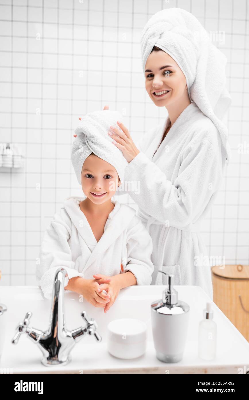 Lächelnde Mutter im Bademantel trägt Handtuch auf dem Kopf des Kindes Im Badezimmer Stockfoto