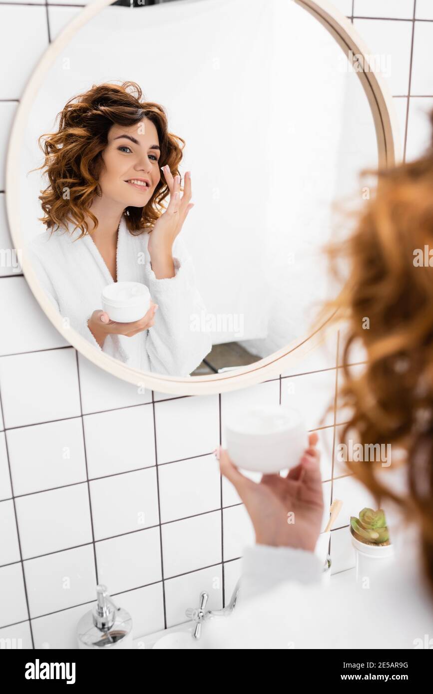 Fröhliche Frau, die kosmetische Creme auf das Gesicht neben Spiegel in Badezimmer Stockfoto
