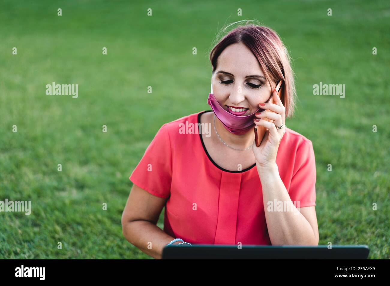 Frau 40 Jahre alt arbeitet im Freien mit Laptop während Coronavirus Outbreak - lächelnder Unternehmer sitzt auf Rasen mit PC Beim Anruf Stockfoto