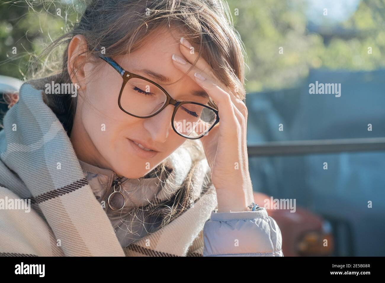 Kopfschmerzen, Migräne, Bluthochdruck. Junge Erwachsene Frau in einer Brille, die ihren Kopf hält, während sie im Freien sitzt, eingewickelt in ein Karli Stockfoto