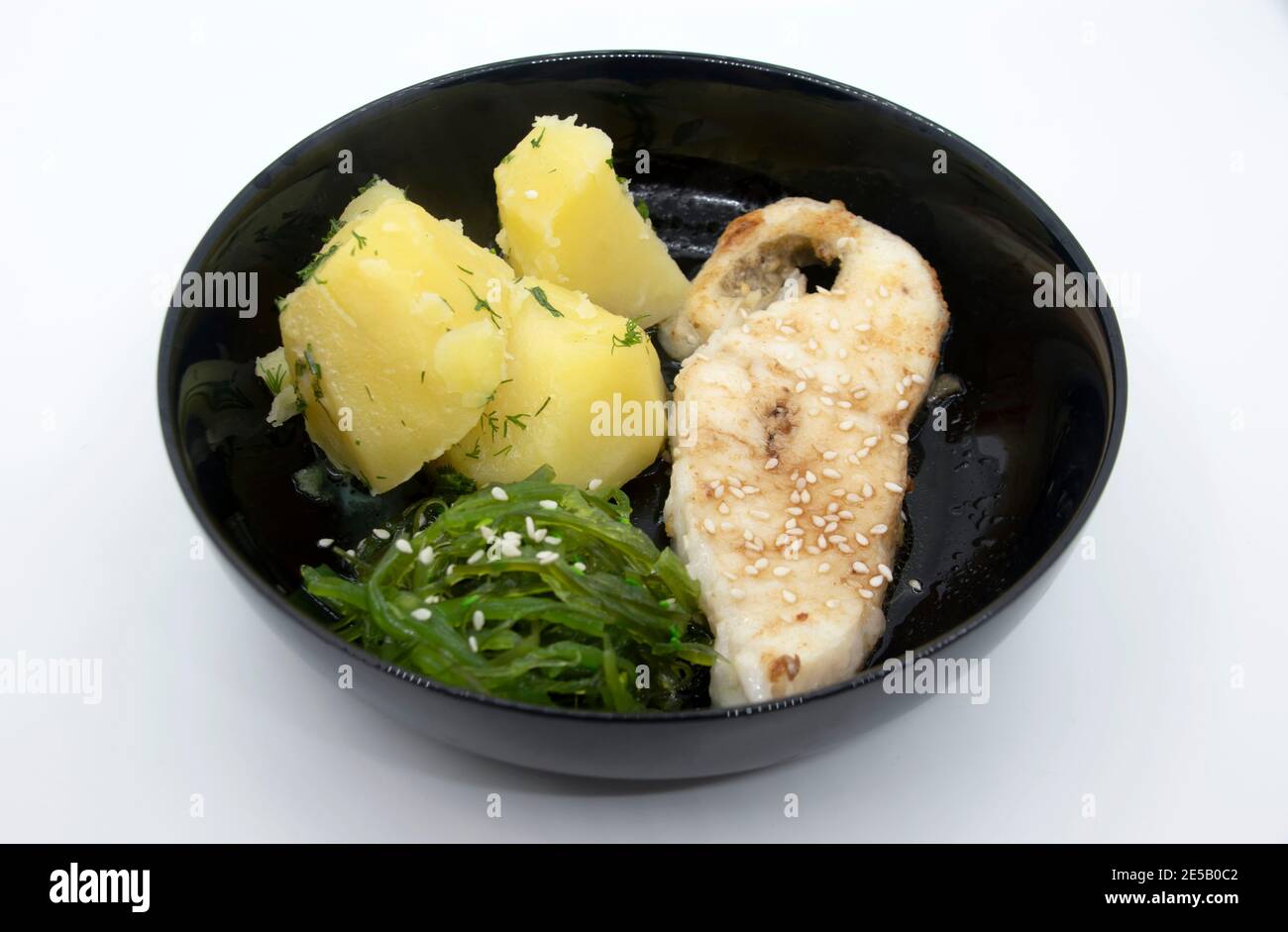 Gebratenes Seefischsteak gekochte Kartoffeln Tschuka Algensalat auf schwarzem Teller. Stockfoto