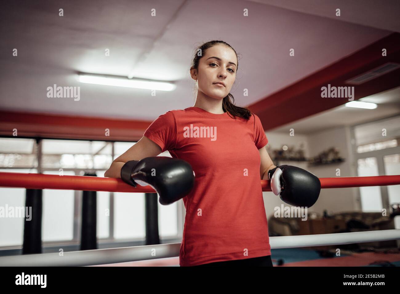 Ein Porträt einer jungen Frau, die sich an einen Boxring lehnt. Eine Sportlerin, die Boxhandschuhe in einem Fitnessstudio trägt. Stockfoto