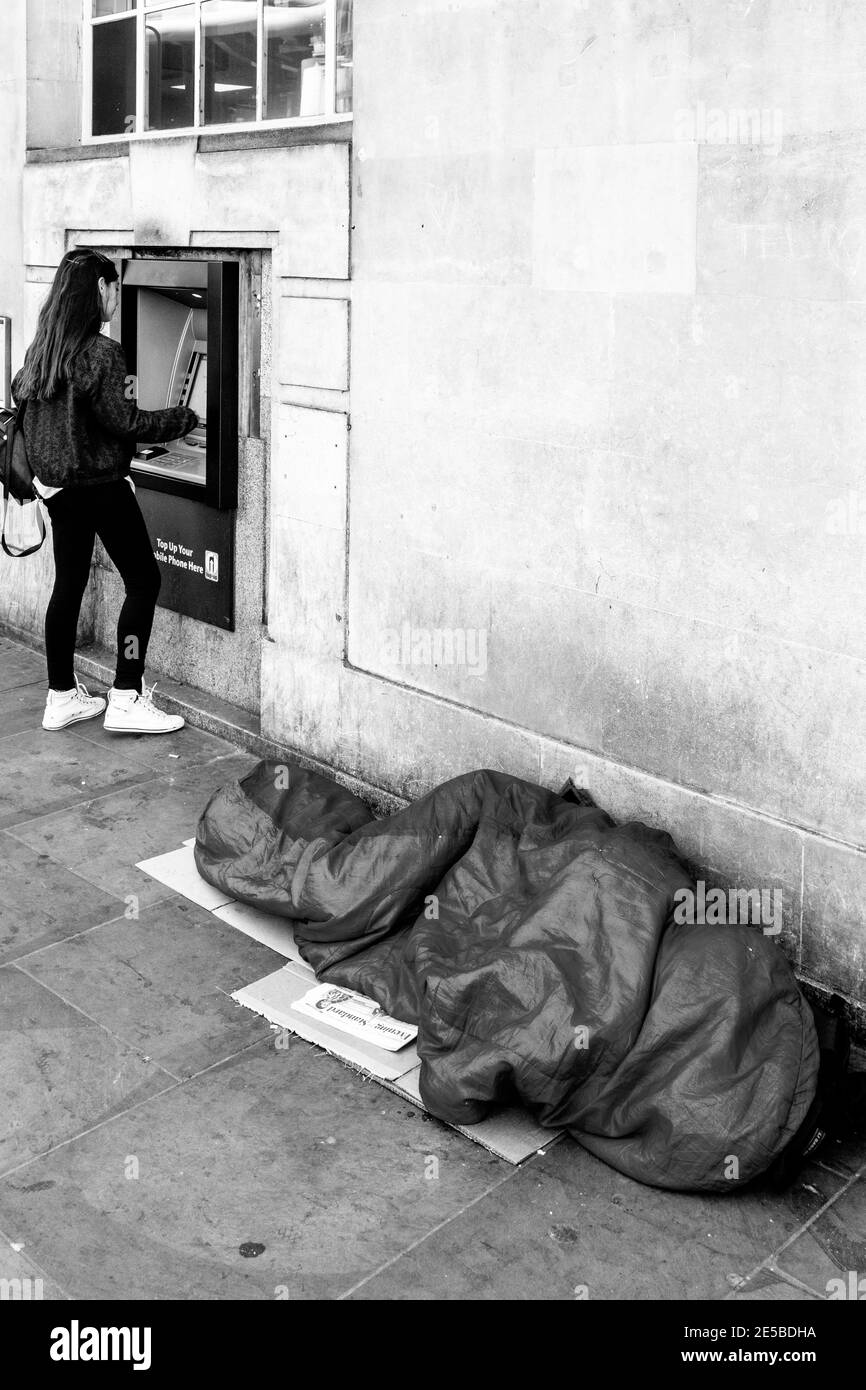 Eine obdachlose Person schläft in EINEM Schlafsack, während EINE junge Frau Geld an einem Geldautomaten in der Borough High Street, London, England abzieht. Stockfoto