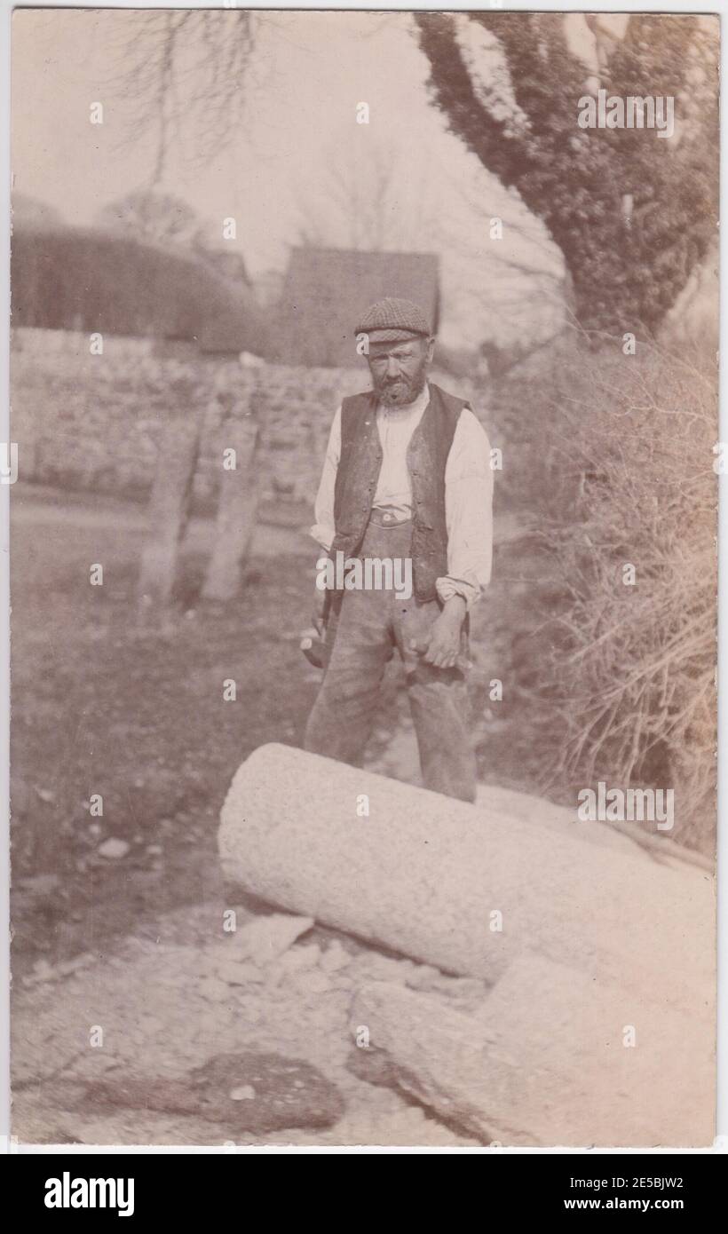 Steinmetz steht mit Hammer und Meißel in den Händen, neben einem runden Steinblock. Grabsteine sind im Hintergrund zu sehen, 1900er / 1910er Stockfoto