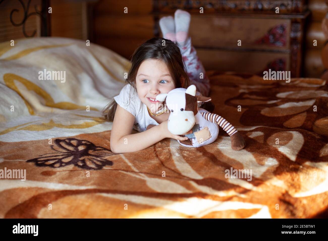Ein fröhliches, ausgesprochenes Mädchen hält eine Spielzeugkuh und liegt an einem sonnigen Morgen auf einem Bett im Schlafzimmer eines rustikalen Blockhauses Stockfoto