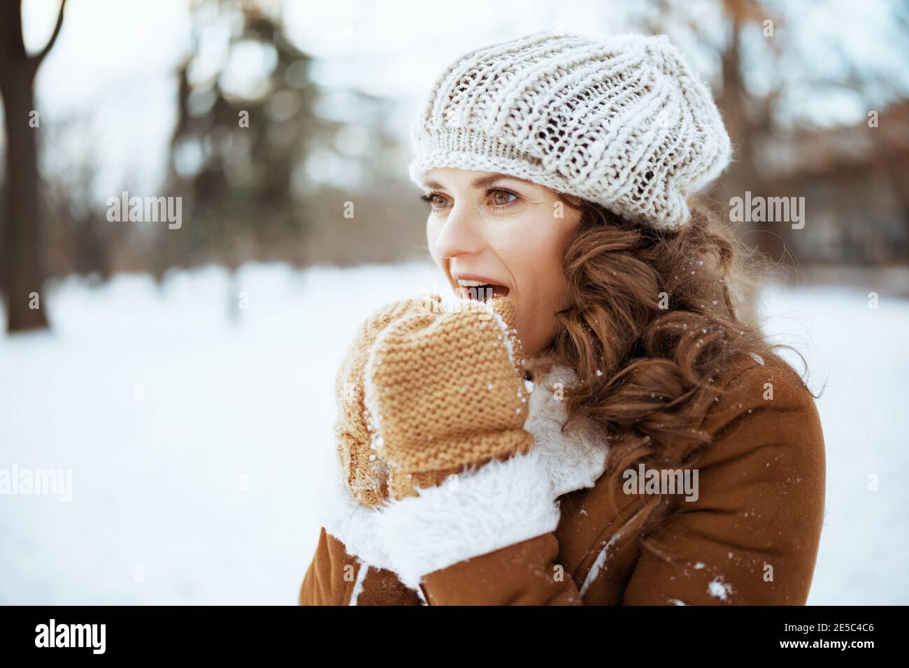 Moderne Frau mittleren Alters mit Fäustlingen in einem gestrickten Hut und Schaffell Mantel wärmende Hände mit Atem im Freien im Stadtpark im Winter. Stockfoto
