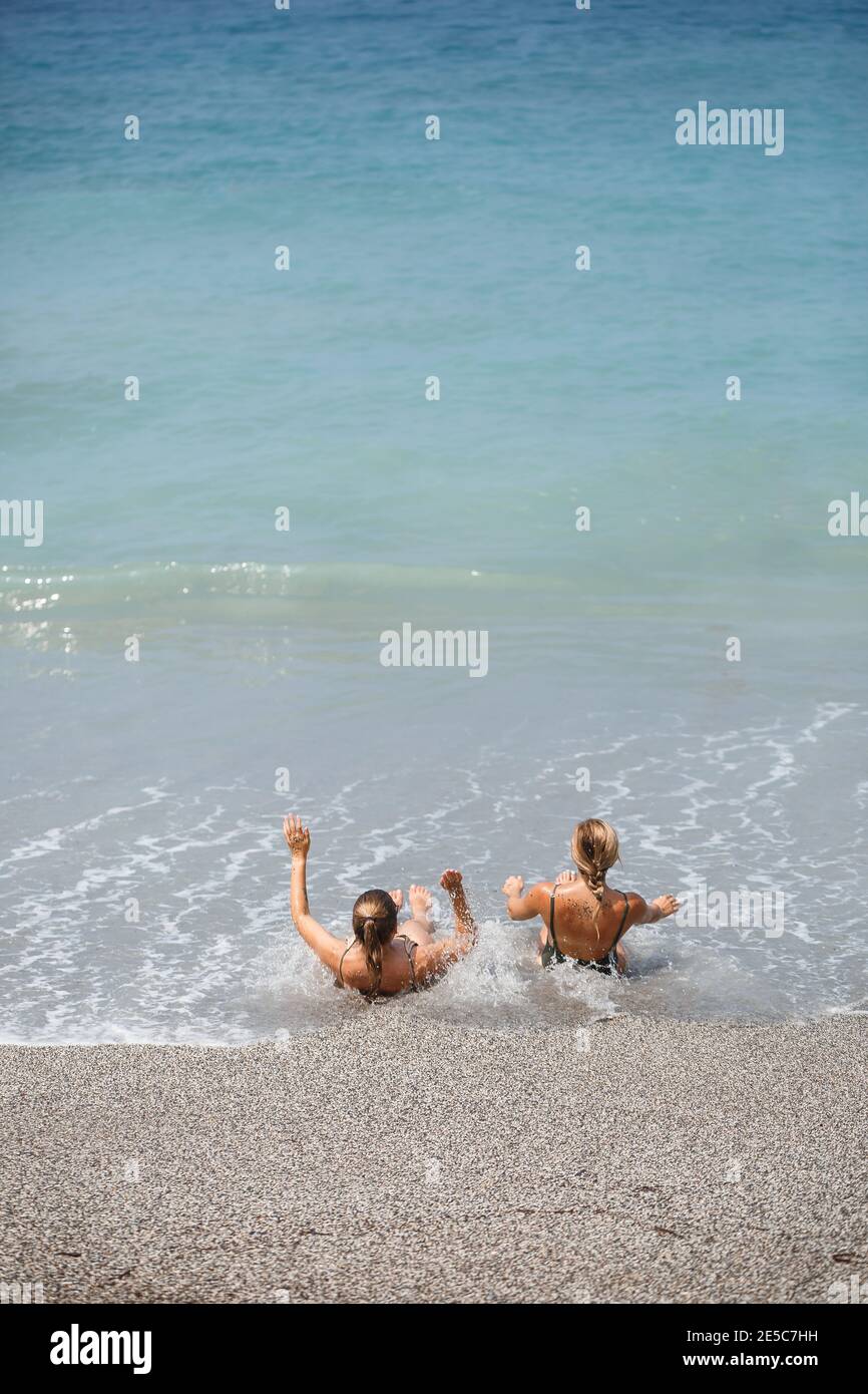 Zwei Mädchen Freundin sitzen auf dem Meer Sandstrand Und die Wellen tränkten sie in Badeanzügen auf einem Sonniger, warmer Tag Stockfoto