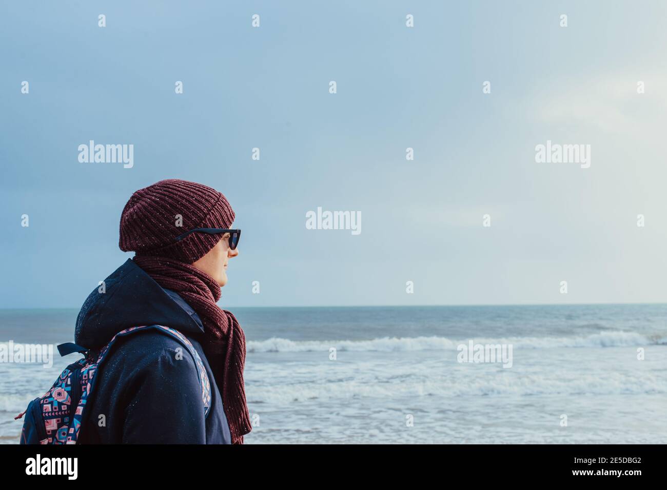 Ein junger Mann in warmen Kleidern und Sonnenbrillen, der am Winterufer spazierengeht und den Moment genießt. Entspannen Sie sich bei einem Spaziergang an der Küste. Selbstreisen vor Ort Stockfoto
