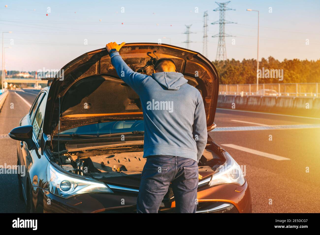 Mann mit seinem kaputten Auto, der den Motor am Straßenrand der Autobahn bei Sonnenuntergang überprüft. Auto bricht auf der Autobahn ab. Öffnen Sie die Motorhaube Stockfoto