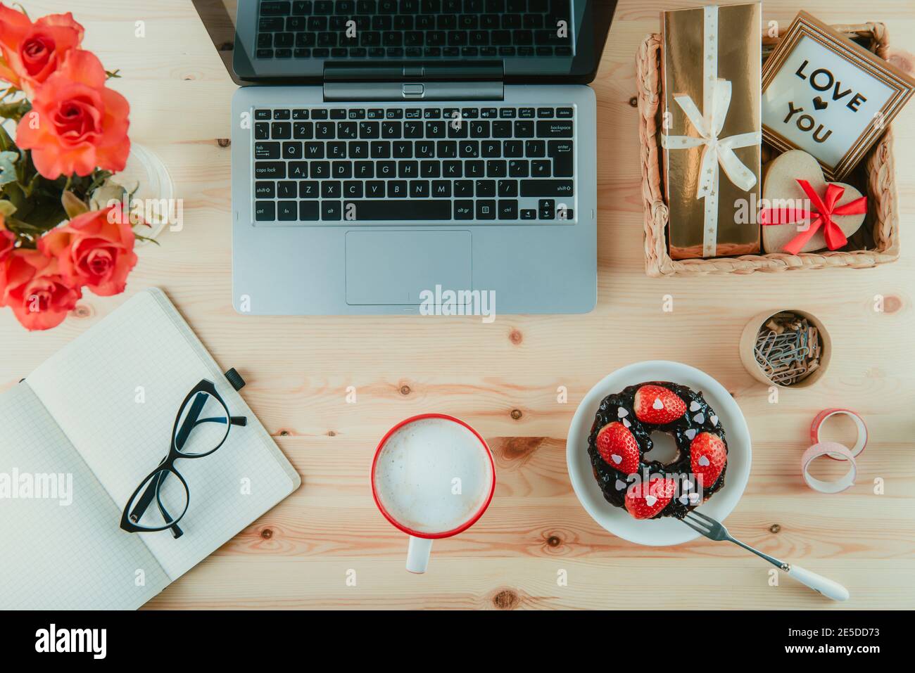 Arbeitsplatz von oben mit Laptop, Notebook, Brille und süßem Valentinstag-Überraschungsfrühstück. Tasse Latte Kaffee, Bagel mit Erdbeeren und Stockfoto