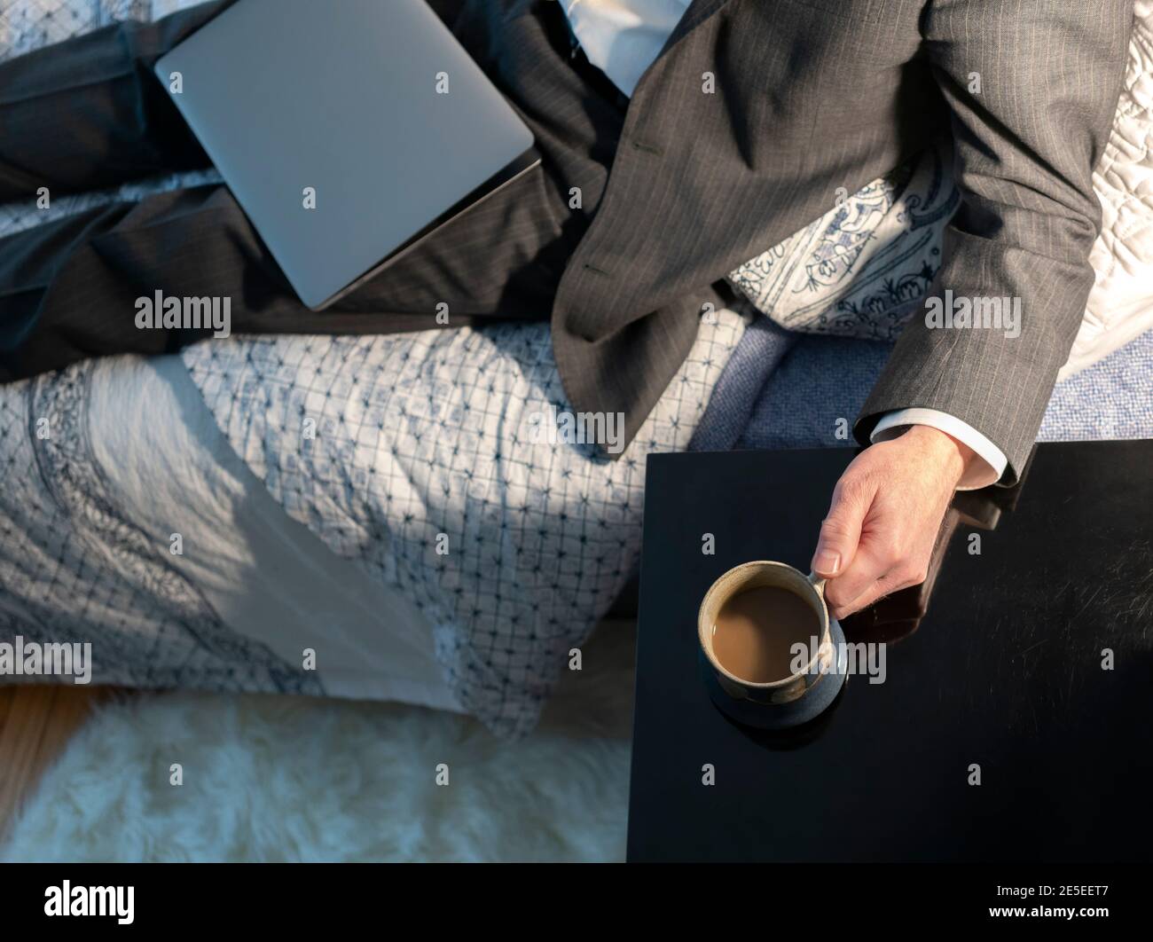 Kaffeepause Arbeit von zu Hause Mann in einem Anzug geschlossenen Laptop auf dem Bett Hand auf Kaffeetasse, Zeitgenössische Coronavirus Arbeit von zu Hause Konzept MR Stockfoto