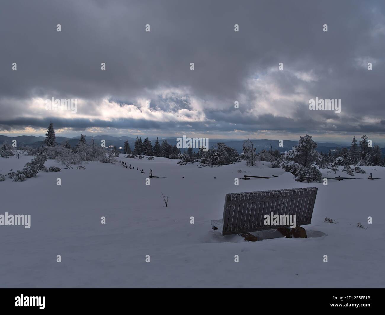 Atemberaubende Winterlandschaft mit Holzbank und Zaun im Tiefschnee, gefrorenen Bäumen und Panoramablick über die Schwarzwälder Hügel am Horizont. Stockfoto