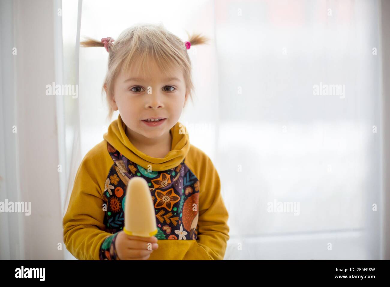 Niedliches süßes Kind, Kleinkind mit kleinen Pferdeschwänzen, zu Hause goofying, Eis essen, lustige Gesichter machen und lächeln Stockfoto
