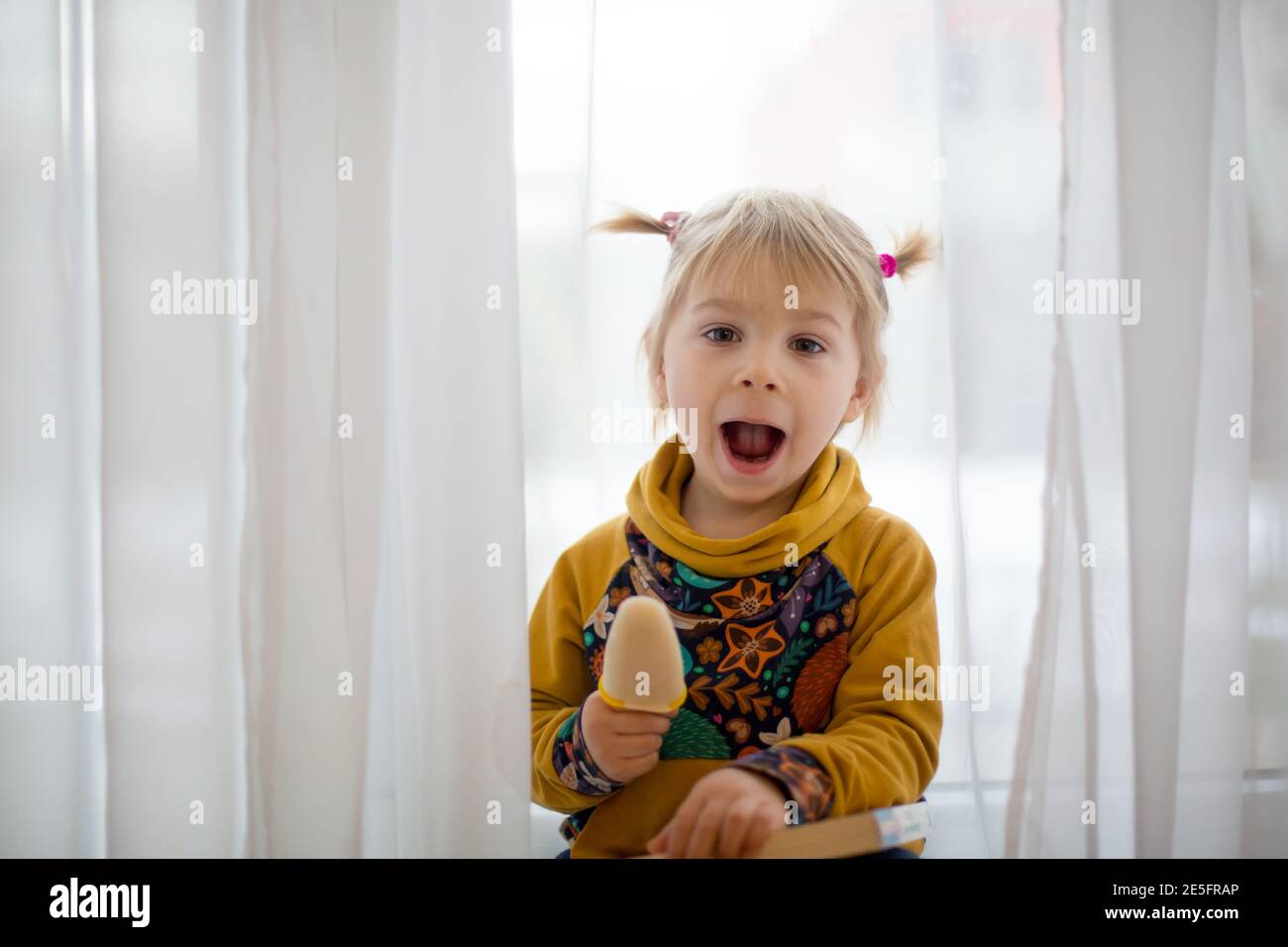 Niedliches süßes Kind, Kleinkind mit kleinen Pferdeschwänzen, zu Hause goofying, Eis essen, lustige Gesichter machen und lächeln Stockfoto