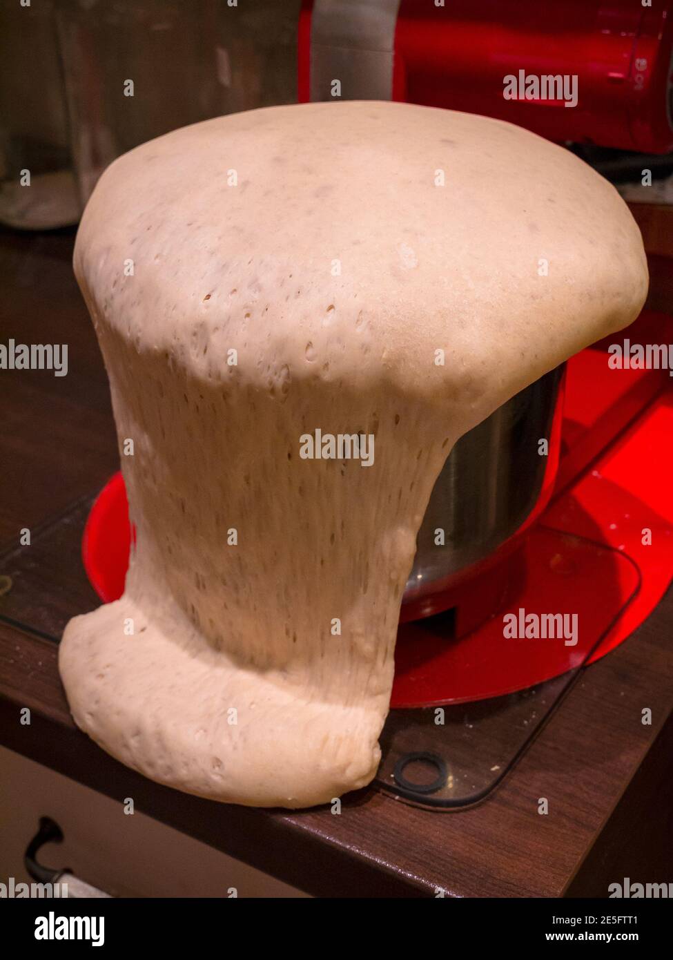 Teig überläuft aus der Schüssel, die auf Glasmatte auf Holztisch steht. Im Hintergrund ist der Küchenroboter und das rote Pad zu sehen. Stockfoto