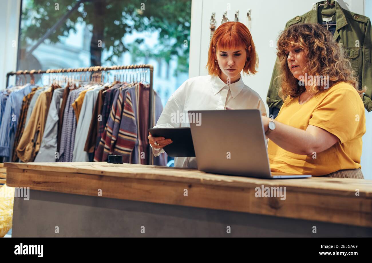 Zwei Frauen, die im Bekleidungsgeschäft arbeiten, suchen an der Theke einen Laptop. Partner arbeiten zusammen im Bekleidungsgeschäft. Stockfoto