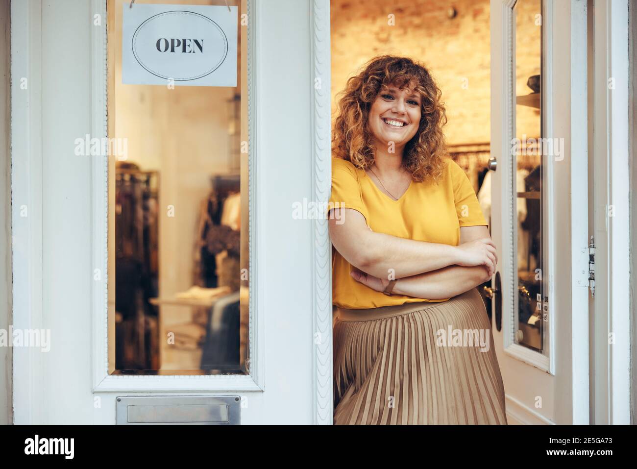 Porträt einer mittleren erwachsenen Frau, die vor der Tür ihrer Modeboutique steht. Erfolgreiche Geschäftsfrau, die vor der Eingangstür ihres Ladens stand und sich ansah, kam Stockfoto