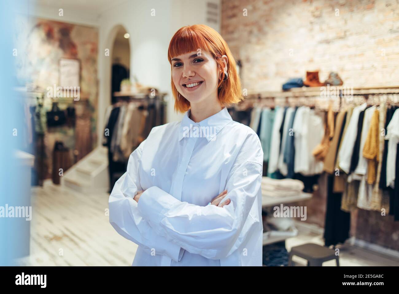 Porträt einer Frau, die in ihrer Boutique steht. Geschäftsfrau, die mit überkreuzten Armen steht und in die Kamera blickt und in ihrem Modegeschäft lächelt. Stockfoto