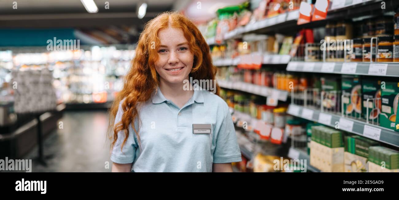 Glückliche junge Verkäuferin im modernen Supermarkt. Frau auf einem Ferienjob im Lebensmittelgeschäft lächelnd an der Kamera. Stockfoto