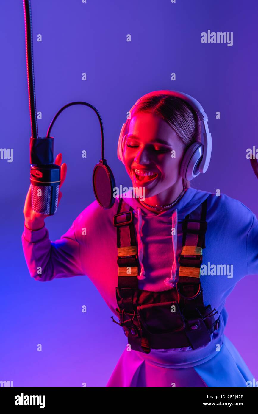 Fröhliche Frau in kabellosen Kopfhörern, die beim Singen Lieder aufzeichnet Mikrofon auf violett mit Farbfilter Stockfoto