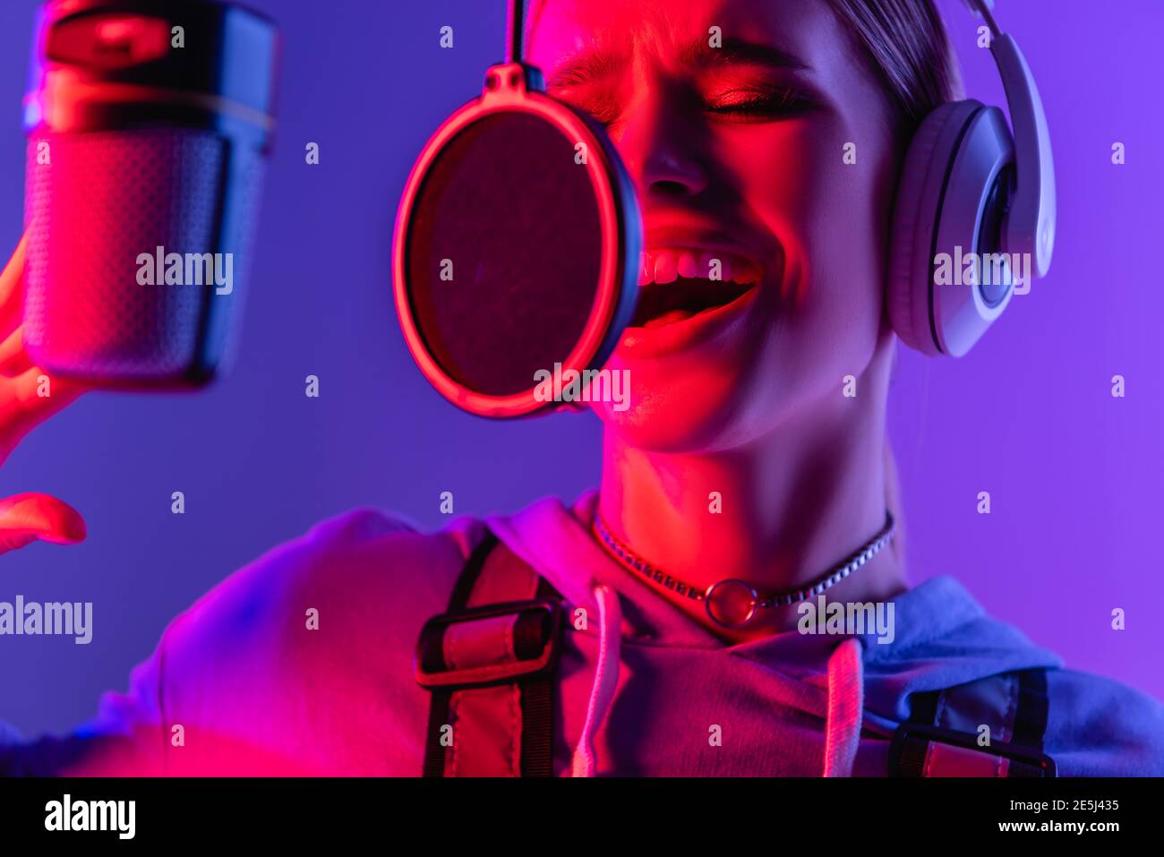 Junge Sängerin in kabellosen Kopfhörern Aufnahme Song während des Singens in Mikrofon auf violett mit Farbfilter Stockfoto