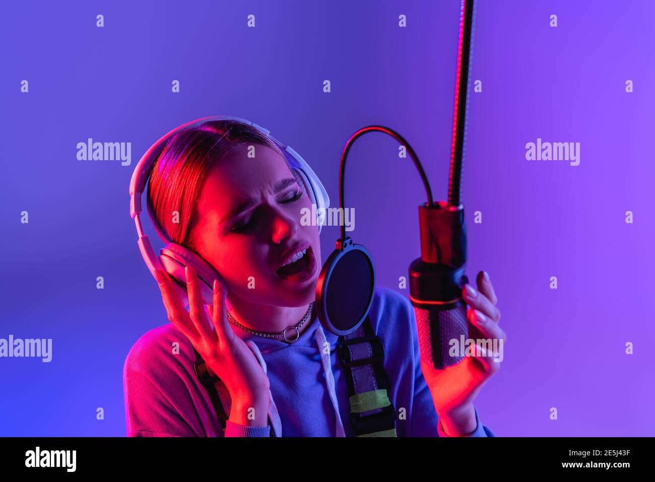 Junge Sängerin in Kopfhörern Aufnahme Song, während im Mikrofon singen Auf violett mit Farbfilter Stockfoto